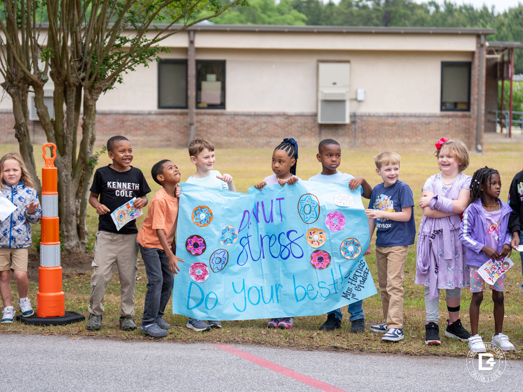 Students stand outside along a school driveway holding handmade signs, including a large blue “Donut stress, do your best” poster decorated with colorful donut drawings.