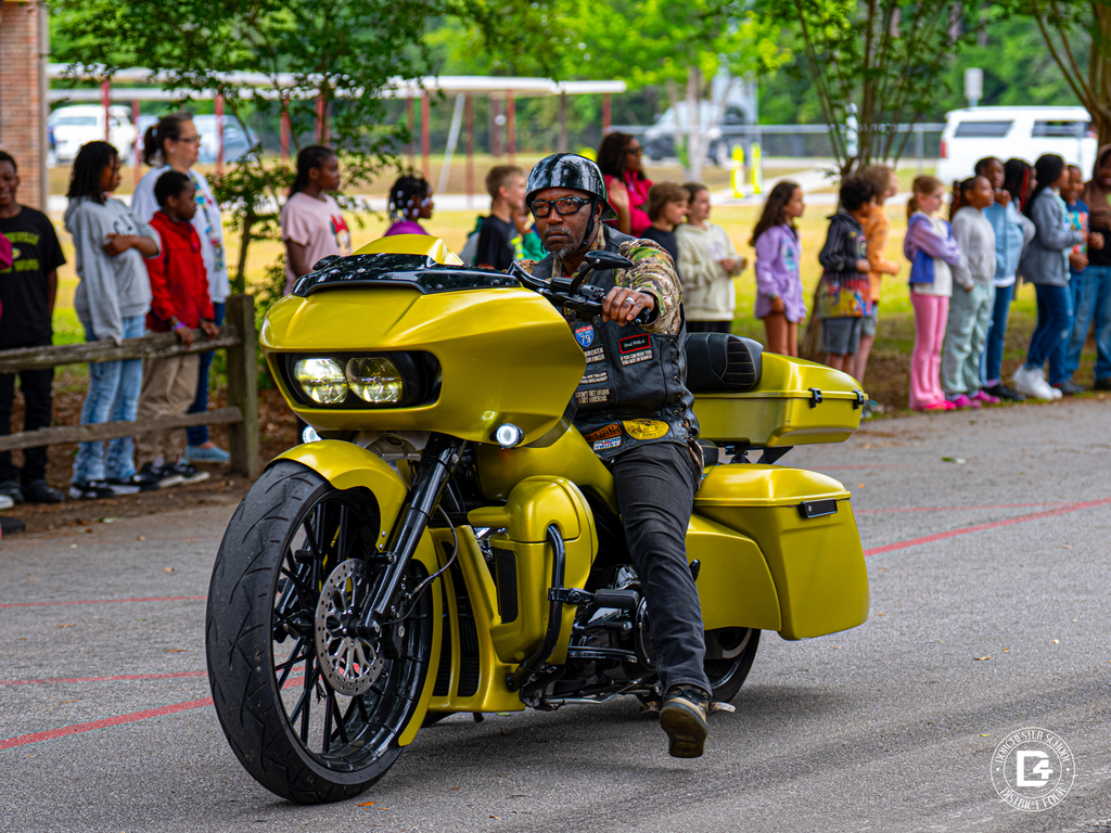 A motorcycle rider on a bright yellow motorcycle passes students lined along the school driveway during the parade.