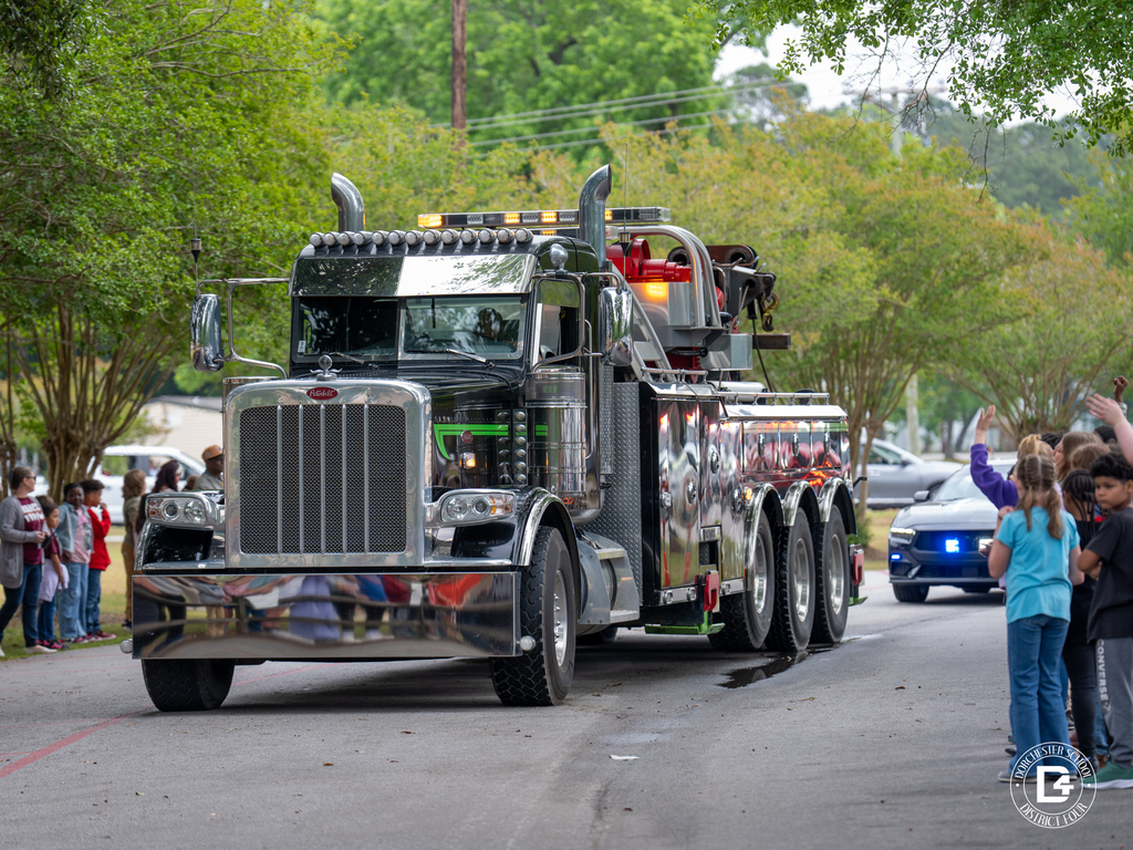 A large tow truck drives through a school parade route as students stand along both sides of the driveway and wave.