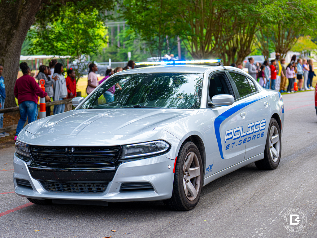 A white St. George police car drives through the school parade route with blue lights flashing while students and staff stand nearby.