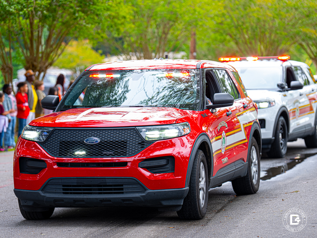 Red and white emergency vehicles drive through the school parade route with lights flashing as students watch from the side of the road.
