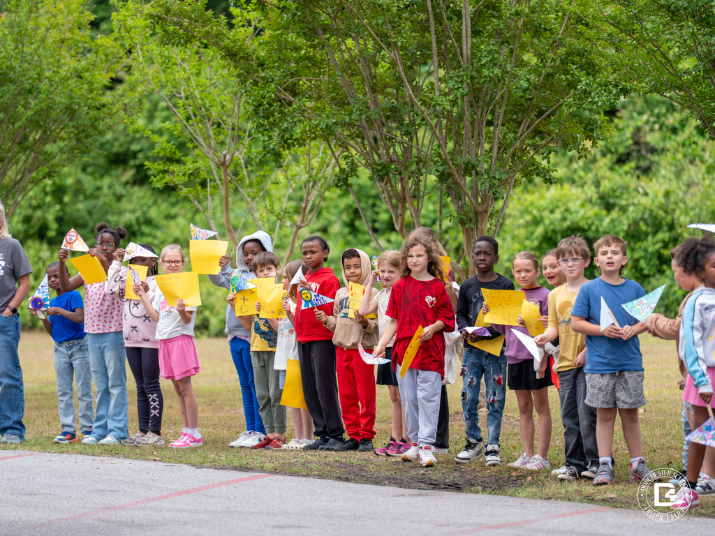 Students line the school driveway holding handmade signs and paper crafts as they watch the parade pass by.