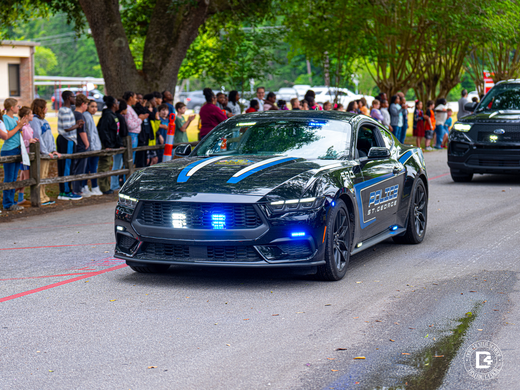 A St. George police vehicle with flashing blue lights drives through the school parade route while students and staff watch from the sidewalk.