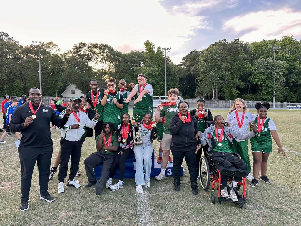 Woodland High School Unified Track & Field team members and coaches pose on the track after winning Lower State, smiling and holding medals, a trophy, and a plaque.