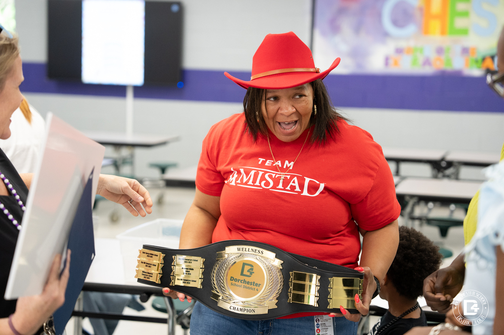 Principal Lemon, wearing a red cowboy hat and “Team Amistad” shirt, proudly holds the Dorchester School District Four wellness championship belt during the Clay Hill Elementary celebration event.