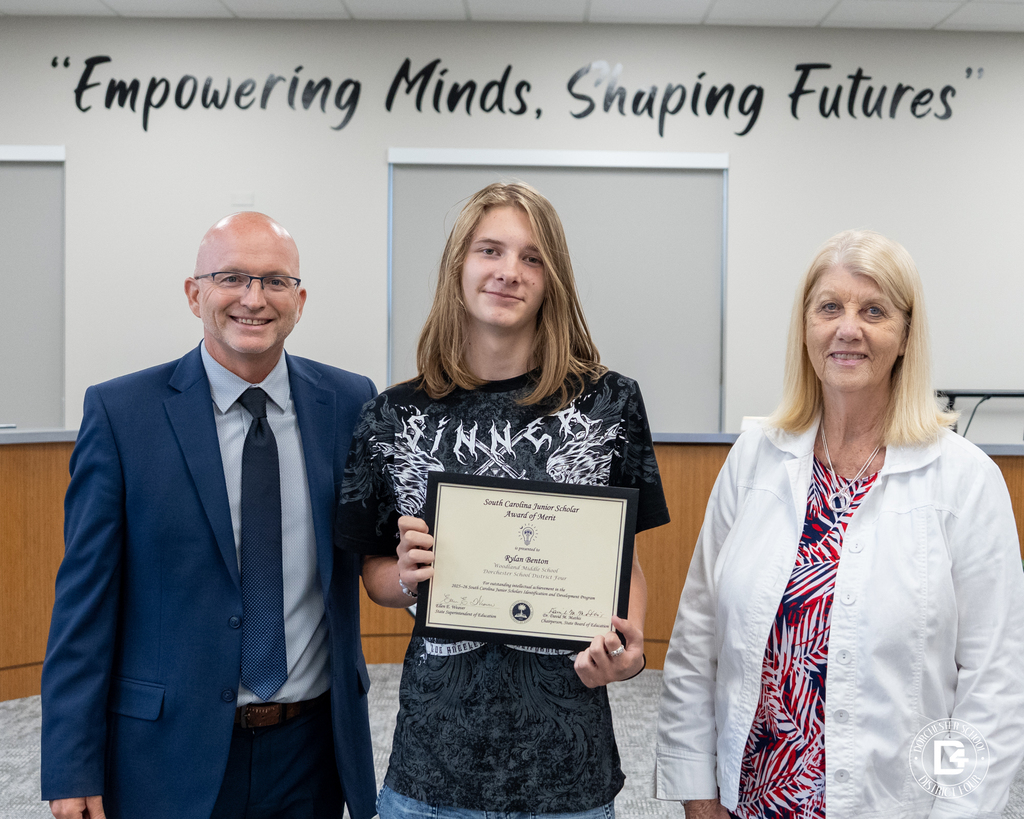 Ryland Benton stands between two district representatives while holding his South Carolina Junior Scholar Award of Merit certificate during the May 19 DD4 Board Meeting.