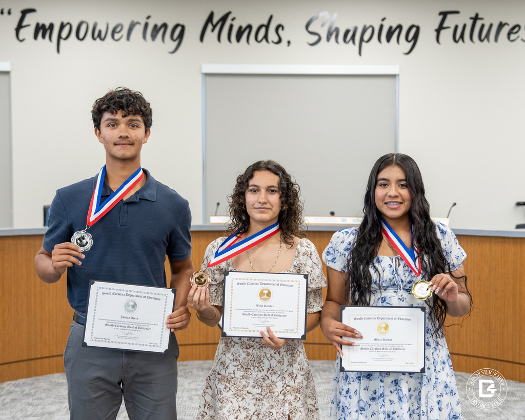 Three students stand together holding their South Carolina Seal of Biliteracy certificates and medals during the May 19 DD4 Board Meeting. From left to right are Joshua Zerra, Bella Benites, and Maria Dávila.