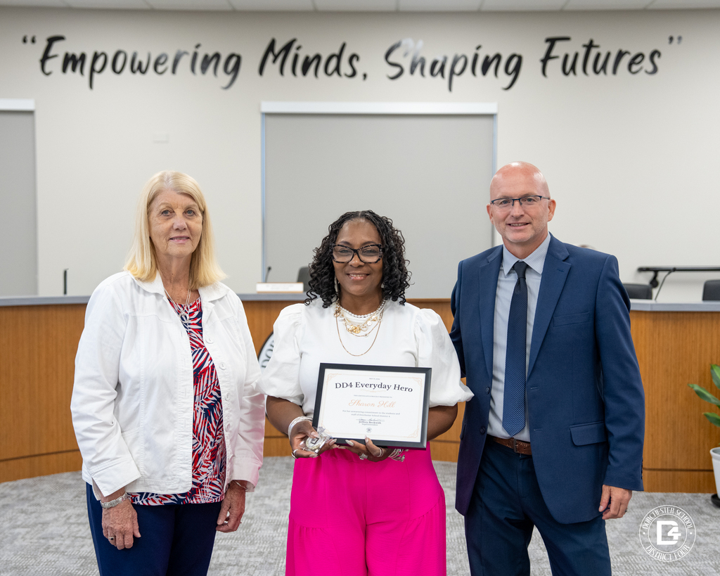 Mrs. Sharon Hill stands between two district representatives while holding her DD4 Everyday Hero award during the May 19 Board Meeting.