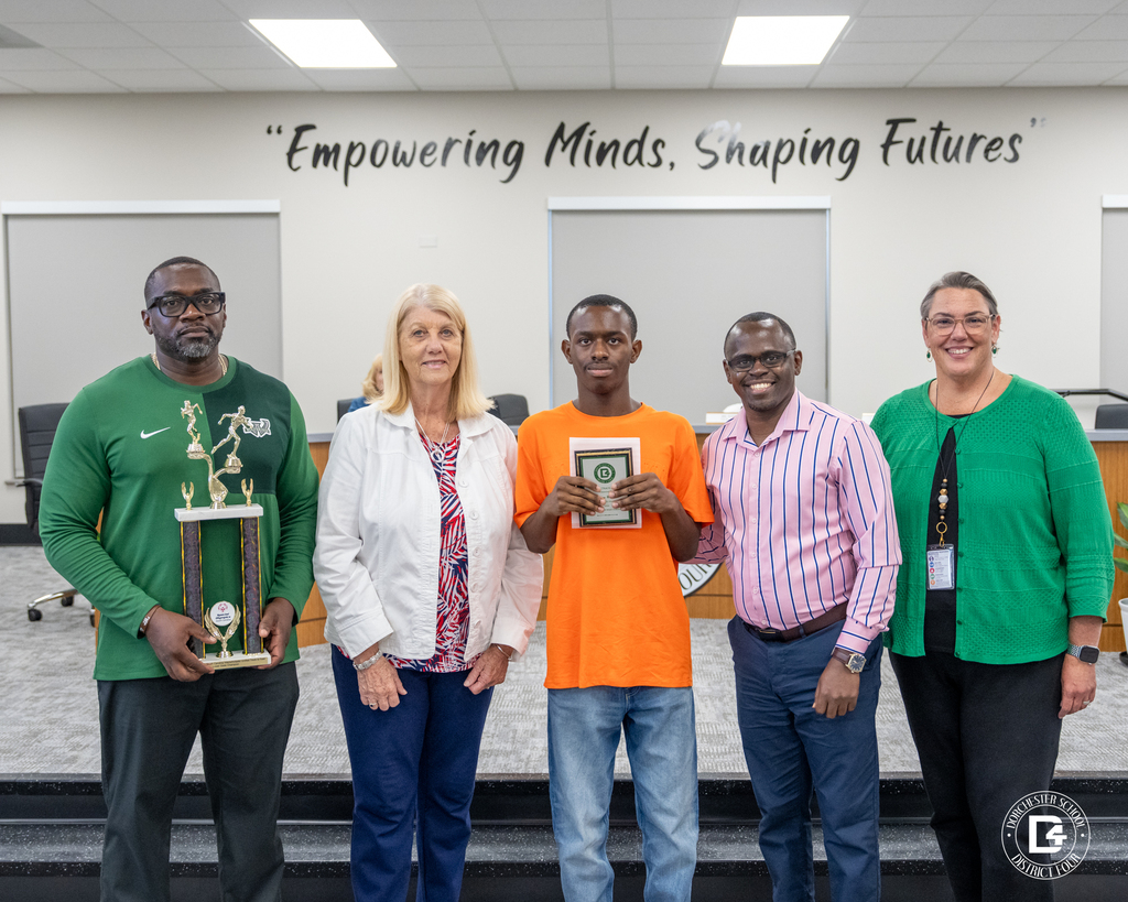 A Unified Track & Field student athlete in an orange shirt stands with coaches and district representatives while holding an award during the May 19 DD4 Board Meeting.