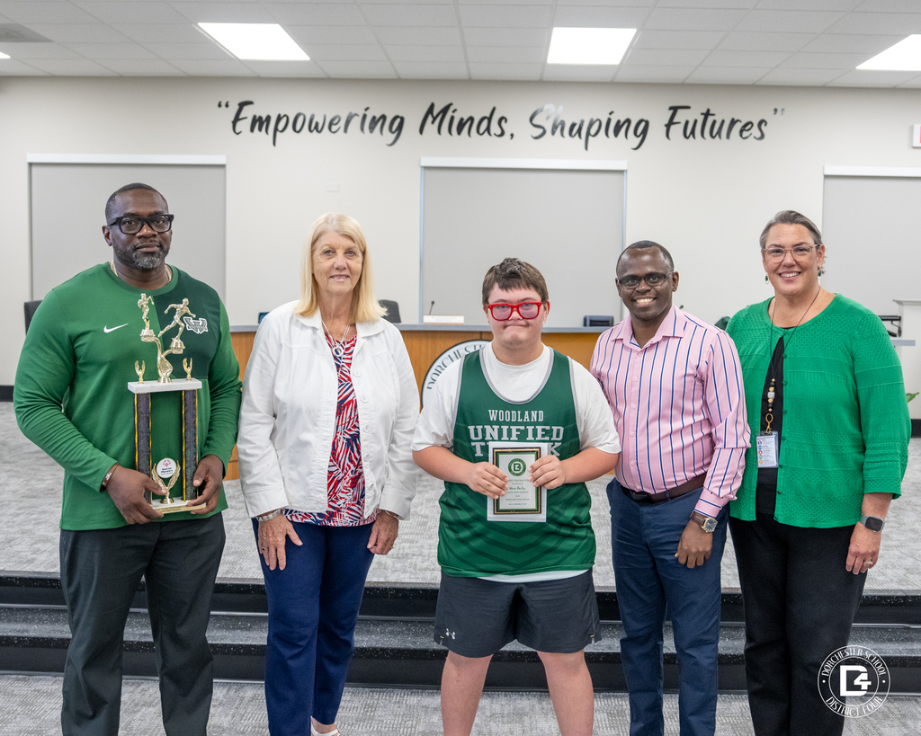 A Unified Track & Field student athlete wearing a Woodland Unified Track jersey stands with coaches and district representatives while holding an award during the May 19 DD4 Board Meeting.