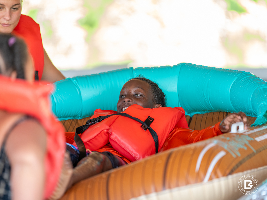 Student wearing a life jacket smiles while riding an inflatable raft through the water safety course.