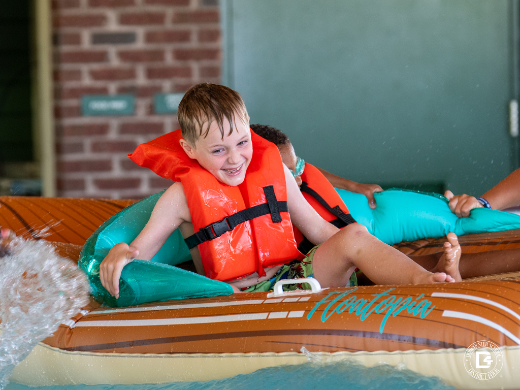Student wearing a life jacket smiles while riding an inflatable raft through the water safety course.