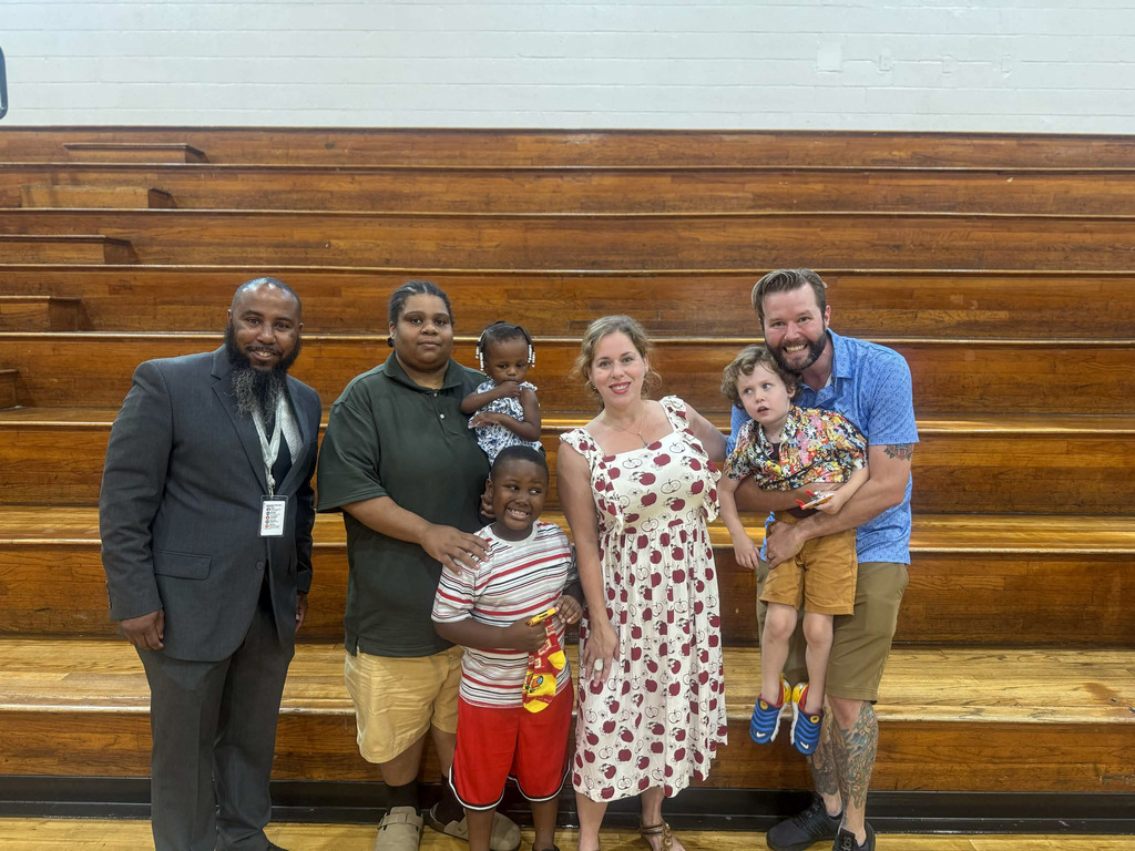A man in a gray suit stands with a family of six in a school gymnasium in front of wooden bleachers. The group includes two adults, a woman in an apple-print dress, a man in a green shirt, and four children, including a young boy in a striped shirt, a toddler girl with hair beads, and a young boy being held by a man in a blue shirt. Everyone is smiling for the photo
