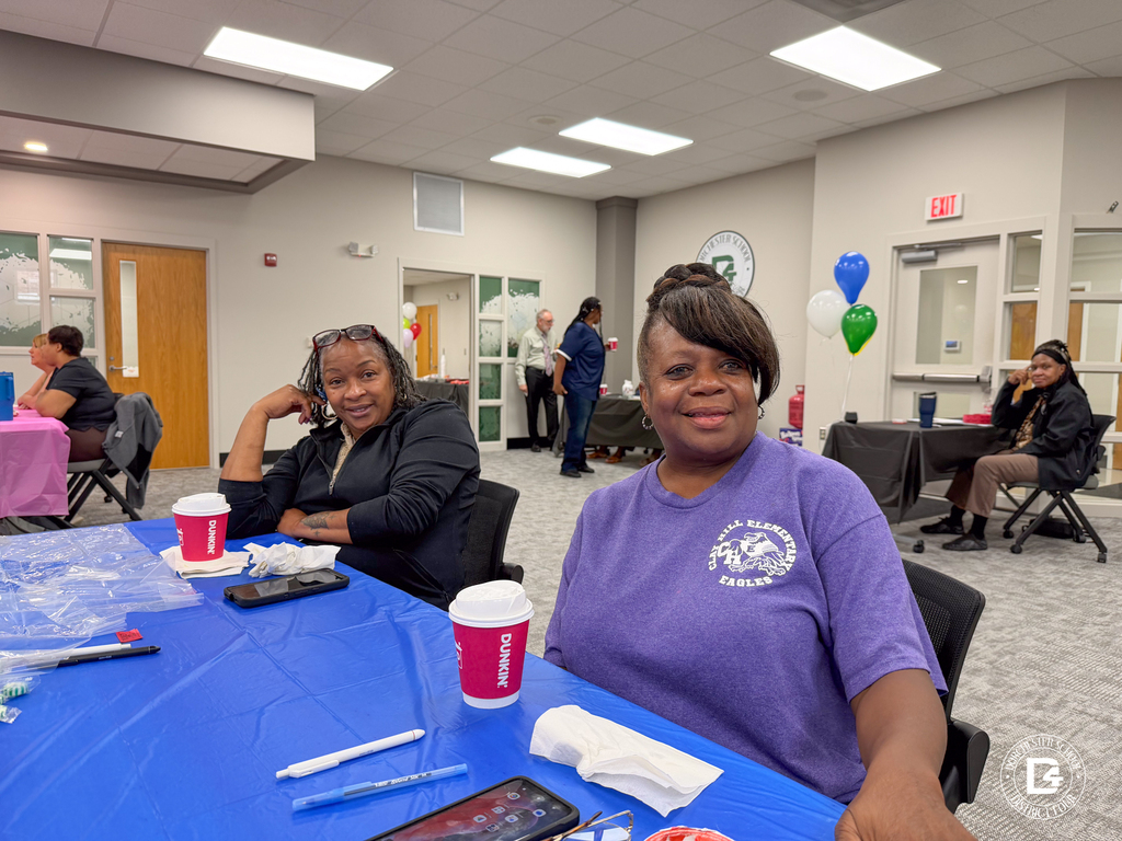 Two DD4 Food Services employees smile at a blue table during the district's Food Services in-service while colleagues gather in the background.