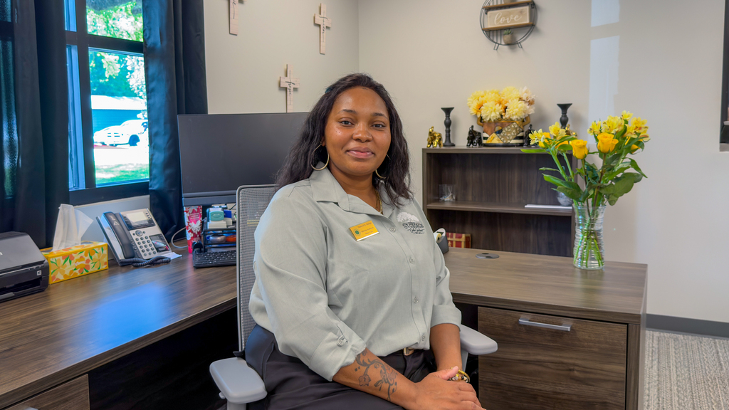 A woman in a light gray Town of St. George uniform shirt seated at a desk in an office decorated with small wooden cross ornaments and a vase of yellow flowers.