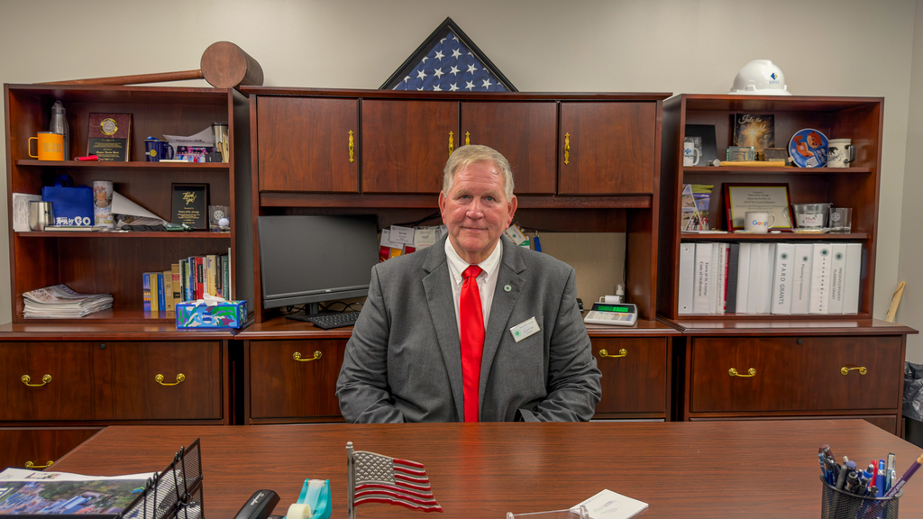 Mayor Kevin Hart seated at a wooden desk in his office, wearing a gray suit and red tie, with bookshelves behind him displaying awards, binders, a folded American flag, and a hard hat.