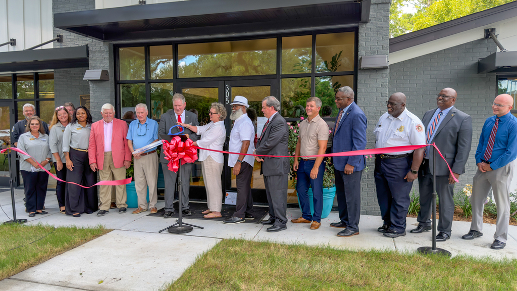 A crowd of local officials and community members gathered outside the Town of St. George's new municipal building for a ribbon cutting ceremony, standing behind a red ribbon with a large bow on a stand. Mayor Kevin Hart stands at the center near the podium.