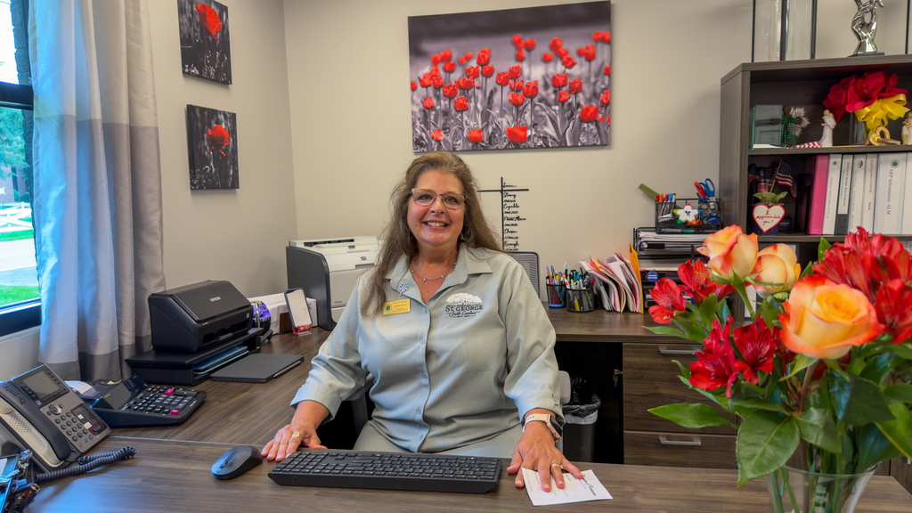 A woman in a light gray Town of St. George uniform shirt seated at a desk in an office decorated with framed poppy photographs and a bouquet of red and orange flowers.