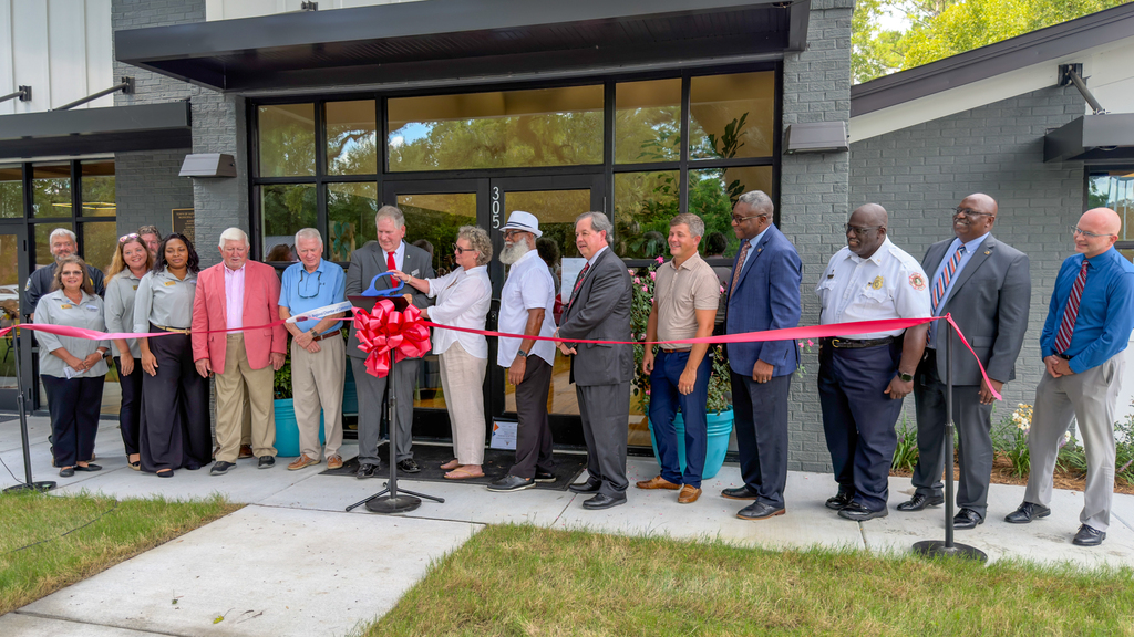 A similar wide shot of the same ribbon cutting ceremony outside the Town of St. George municipal building, with officials lined up behind the red ribbon in front of the entrance marked "305."