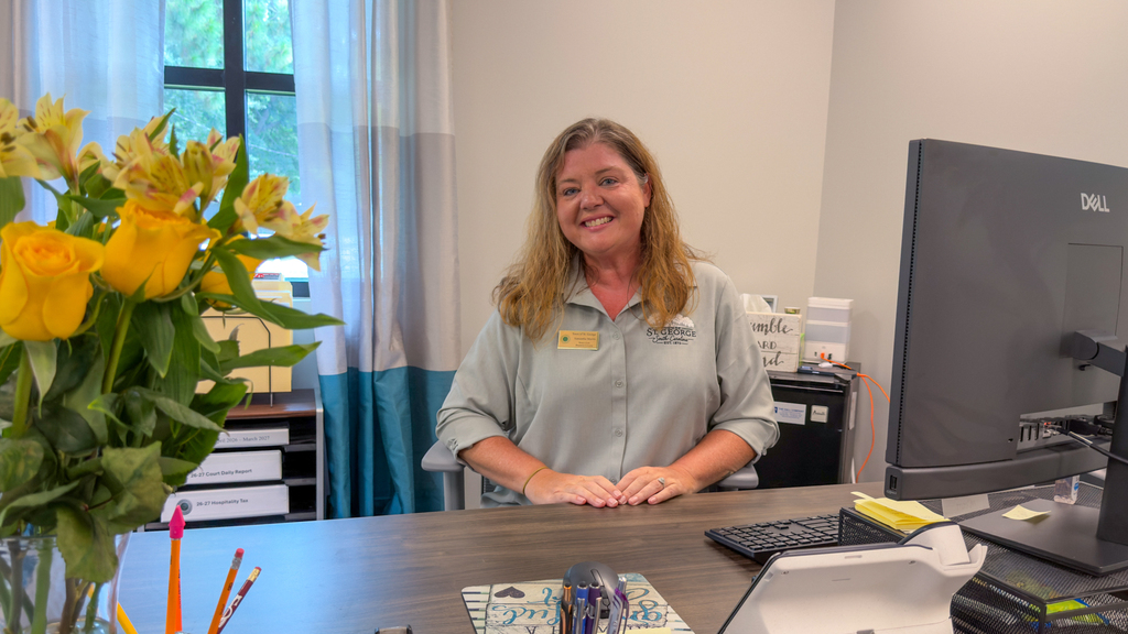 A woman in a light gray Town of St. George uniform shirt standing behind a desk in an office with a Dell monitor, yellow flowers, and decorative signage in the background.