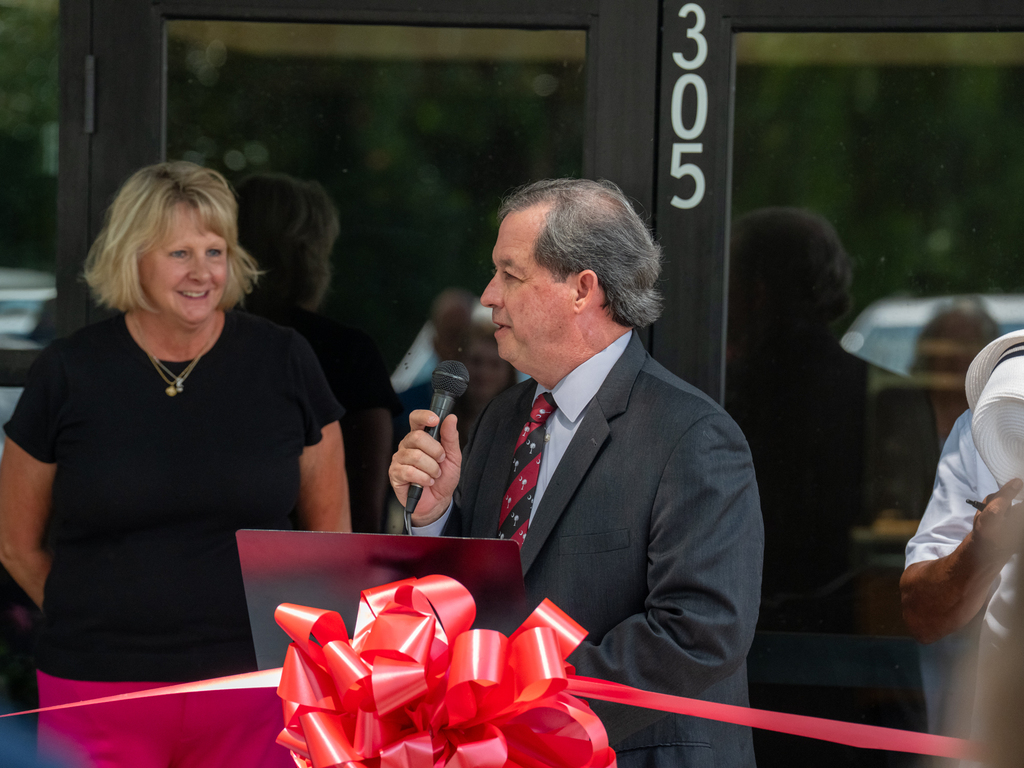 A man in a gray suit speaking into a microphone at a podium decorated with a large red bow, with a woman in a black shirt standing beside him near the building's entrance marked "305."