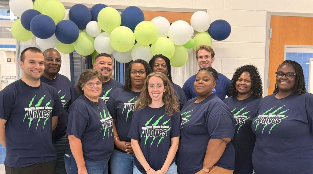 A group of Woodland Middle School staff members wearing matching Wolves shirts pose together beneath an arch of navy, lime green, and white balloons.
