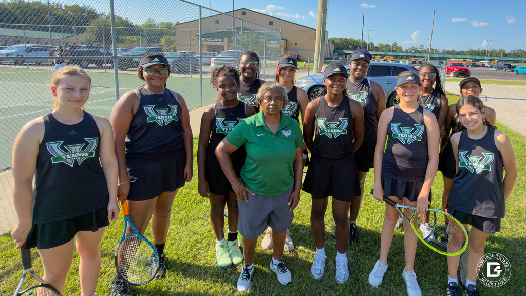 Woodland ladies tennis team posing with their coach on the grass outside of the fencing for the tennis court. . Ladies are positioned in two rows wearing match black uniforms with the woodland logo on the front.