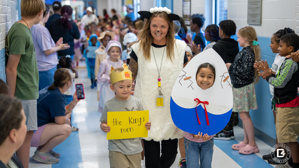 A teacher dressed as Old Mother Hubbard, wearing a bonnet and carrying a cane, walks down the hallway as students in costume follow behind her.