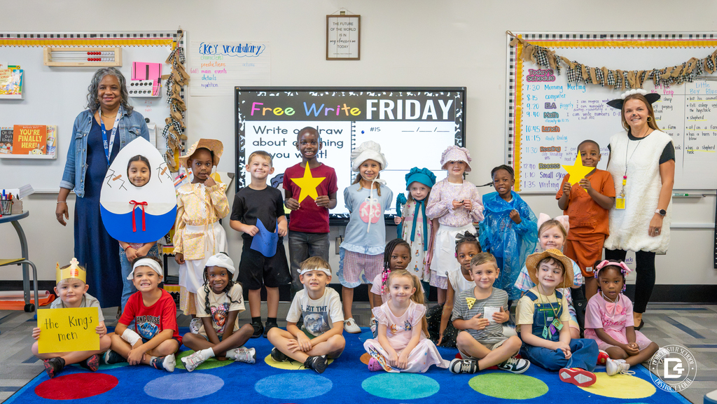 A kindergarten class poses for a group photo in their classroom with two teachers, one dressed as Humpty Dumpty's egg character and one as a sheep, surrounded by students in nursery rhyme costumes.