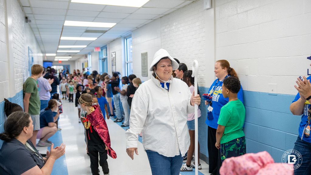A teacher dressed as Old Mother Hubbard, wearing a bonnet and carrying a cane, walks down the hallway as students in costume follow behind her.