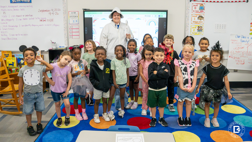 A kindergarten class poses for a group photo on a colorful classroom rug with their teacher dressed as Old Mother Hubbard, students wearing various nursery rhyme costumes including a clock, king, cow, and witch.