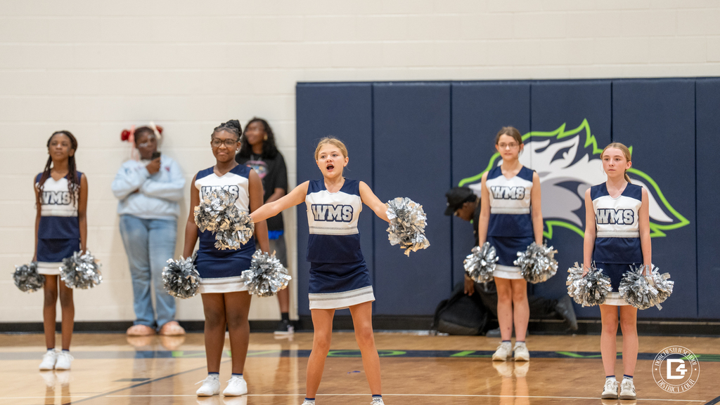 WMS cheerleaders in navy and white uniforms perform with silver pom-poms during the pep rally, one cheerleader leading a chant at center court.