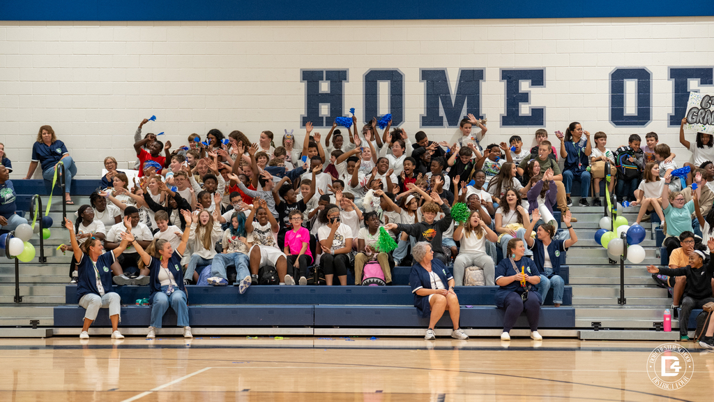 A large group of students and staff in white and navy shirts cheer with their hands raised in the bleachers beneath a "HOME OF" wall sign, with a sign reading "6th grade" visible on the right.