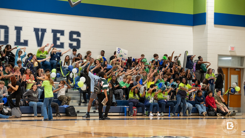 Woodland Middle School students in the bleachers cheer and reach toward the camera, with cheerleaders in yellow and navy pom-poms in the foreground and a sign reading "8th Grade rules! WMS Wolves."