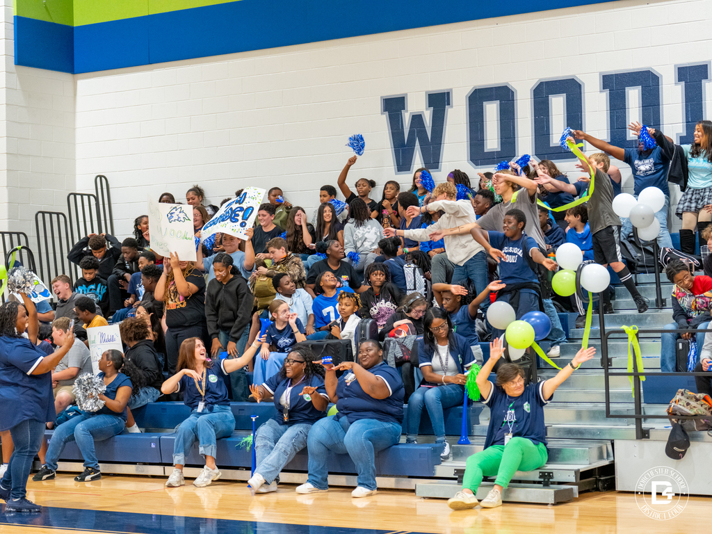 Woodland Middle School staff and students cheer and dance in the bleachers, waving pom-poms and holding signs reading "Go Woodland Wolves" beneath a wall painted with "WOOD."