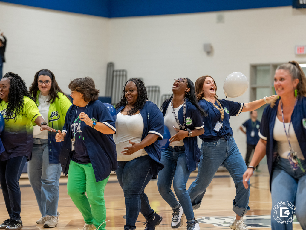 A group of WMS staff members in matching navy jerseys dance and cheer together on the gym floor, one batting a balloon in the air.