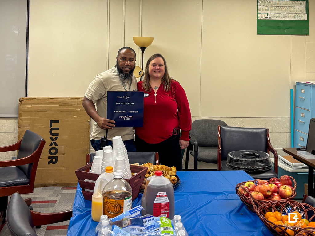 A staff member and a woman wearing a Food Lion name badge smile side by side. He holds a navy sign that reads, "Thank You for All You Do. Breakfast Courtesy Food Lion 2850." In front of them, a table with a blue tablecloth holds stacks of foam cups, a tray of pastries, orange juice, apple juice, tea, bottled water, and baskets of apples and oranges.