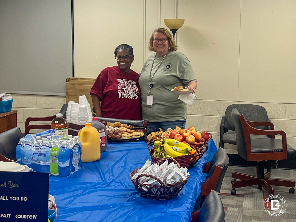 Two smiling staff members stand behind a breakfast table covered with a blue tablecloth. One wears a maroon Tigers T-shirt, and the other wears a green district T-shirt and holds a plate with a pastry. The table is filled with trays of pastries, muffins, and donut holes, baskets of apples, bananas, oranges, and breakfast bars, plus bottled water, orange juice, apple juice, and a jug of tea. Part of a navy thank you sign is visible in the corner.