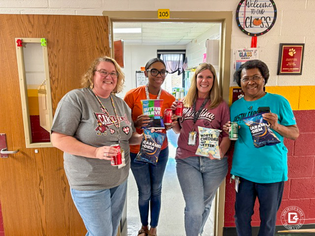 Four smiling Williams Memorial Elementary staff members stand in the doorway of room 105 holding cans of soda and bags of popcorn. Dressed for College Colors Day, they wear a gray Carolina T-shirt, an orange T-shirt, a garnet Carolina T-shirt, and a teal Coastal Carolina T-shirt. Their popcorn bags include Caramel-Diem, White Cheddar Go Getter, and a butter flavor. A "Welcome to Our Class" sign and a framed plaque hang on the wall beside them.