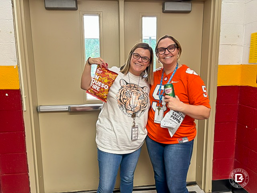 Two smiling staff members pose together in front of a set of double doors in a hallway with maroon and gold painted walls. One wears a cream T-shirt with a tiger face on it and holds up a bag of In Queso Fire popcorn. The other wears an orange Clemson football jersey and holds a can of Canada Dry ginger ale and a bag of salt and pepper popcorn.