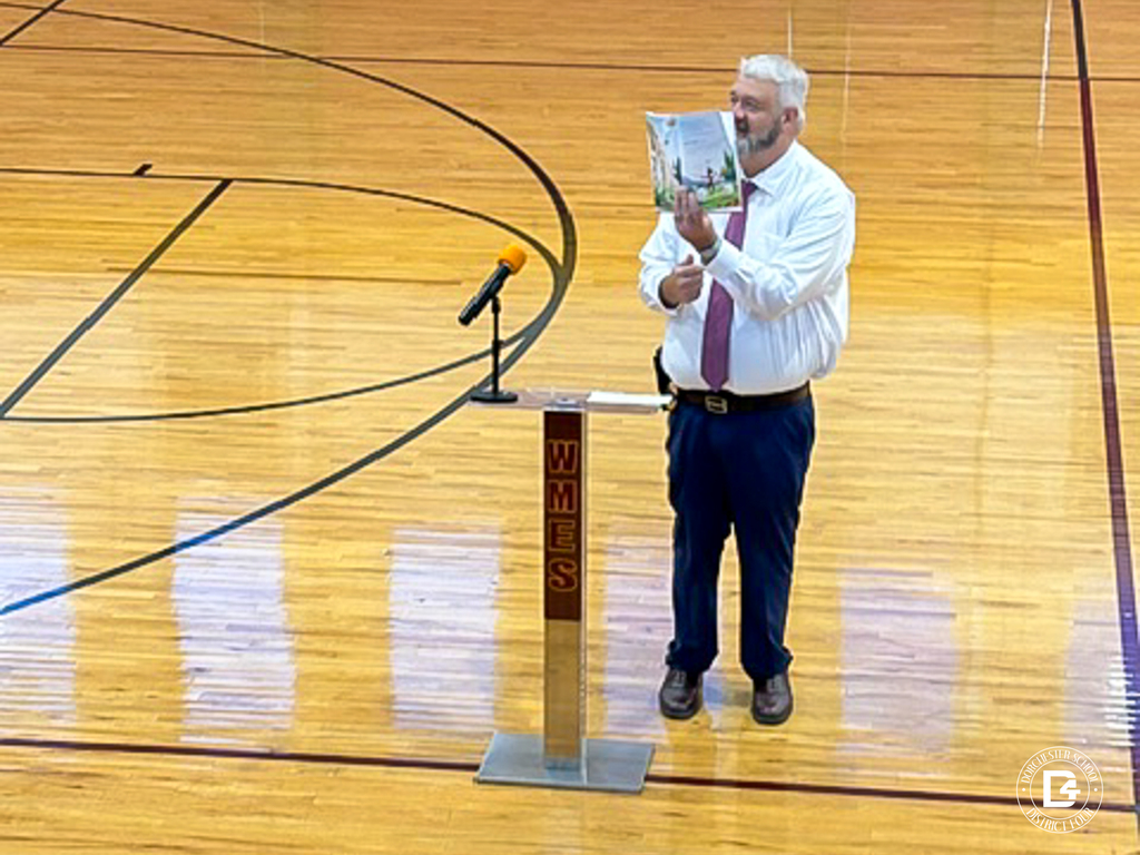 Chief Dunaway stands on the gym floor at Williams Memorial Elementary, holding up an open, colorful picture book for students to see. He wears a white dress shirt, maroon tie, and navy pants and stands beside a clear podium with a maroon WMES sign and a microphone.
