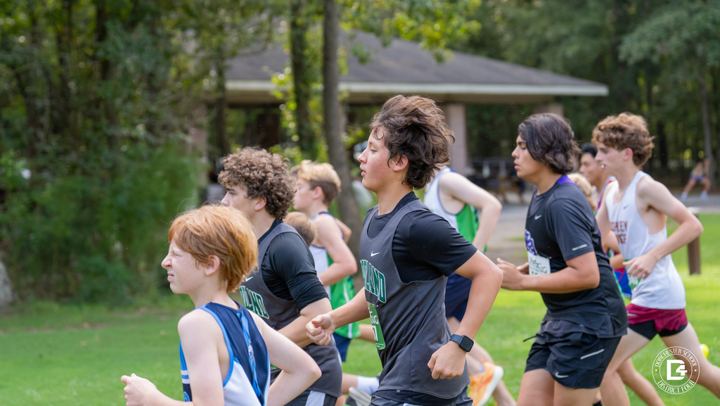A Woodland High School cross country runner in a dark jersey and smartwatch stays focused mid-race at Wannamaker Park, surrounded by runners from other schools with a park pavilion behind them.