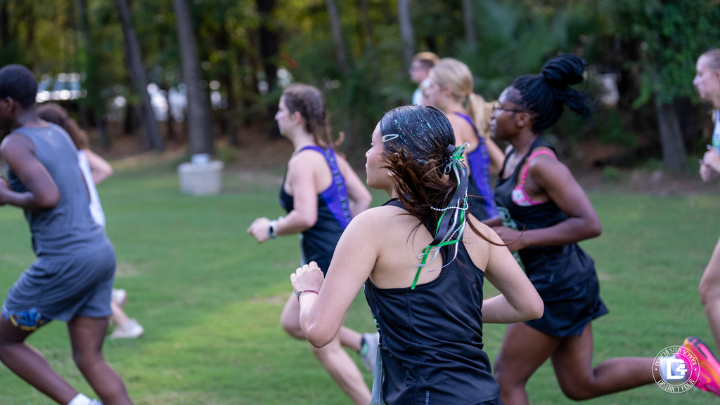 Viewed from behind, a Woodland High School cross country runner with glitter in her hair and green and white ribbons in her ponytail runs in a pack of girls across the grass at Wannamaker Park.