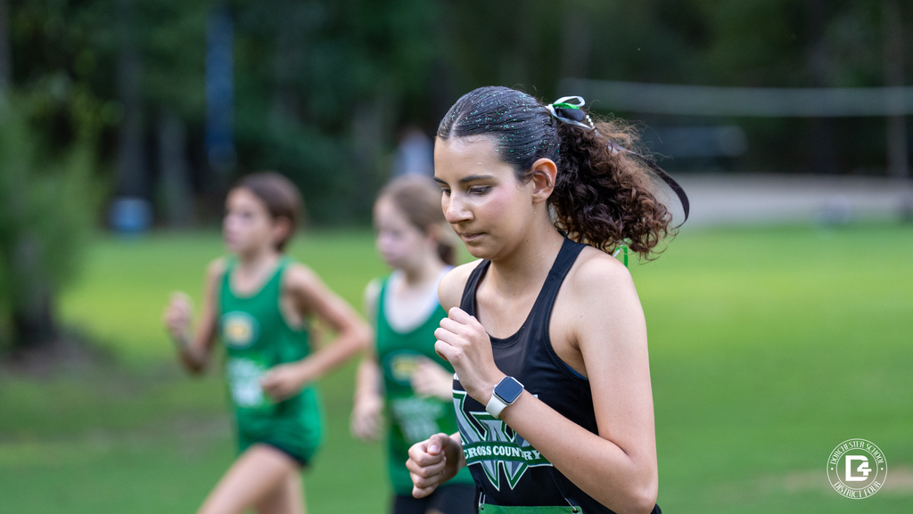 A Woodland High School cross country runner in a dark jersey and smartwatch stays focused mid-race at Wannamaker Park, surrounded by runners from other schools with a park pavilion behind them.