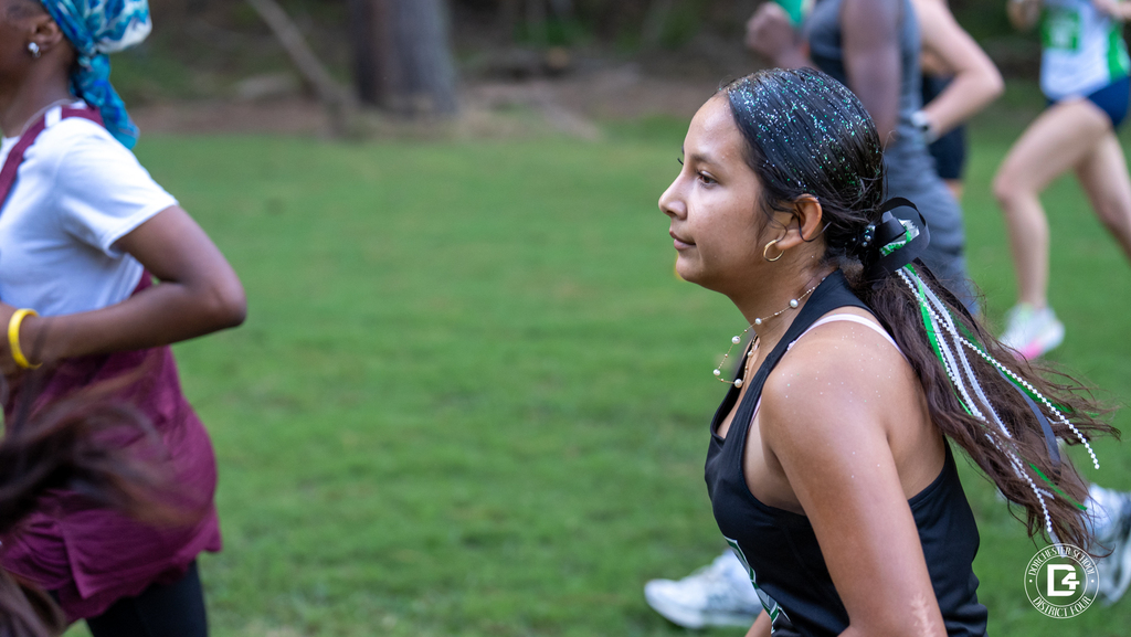 Side view of a Woodland High School cross country runner at Wannamaker Park, with green glitter in her hair, a pearl necklace, and green, white, and black ribbons flowing from her ponytail as she runs.