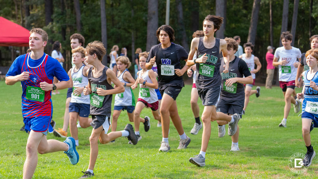 Woodland High School cross country runners wearing bibs 1958, 1954, and 1955 run with athletes from other schools across a grassy field at Wannamaker Park, with tall pine trees behind them.