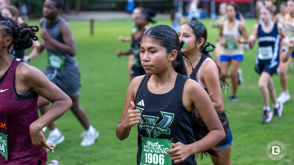 A Woodland High School cross country runner wearing bib 1906 and green glitter in her hair runs with determination in a pack of girls at Wannamaker Park, with a teammate close behind.