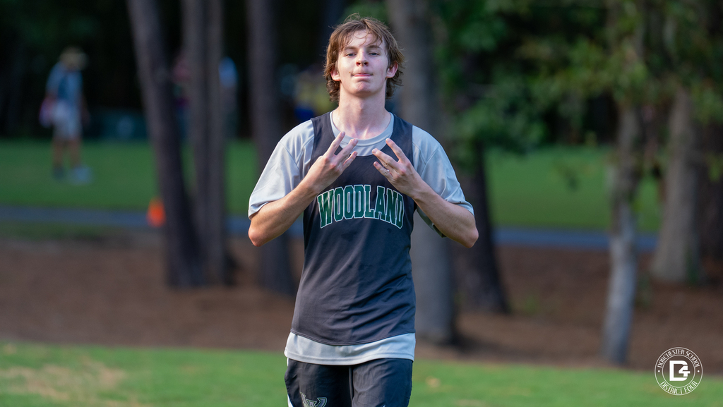 A Woodland High School cross country runner in a black Woodland jersey over a gray shirt walks toward the camera at Wannamaker Park, holding up both hands in a W sign.