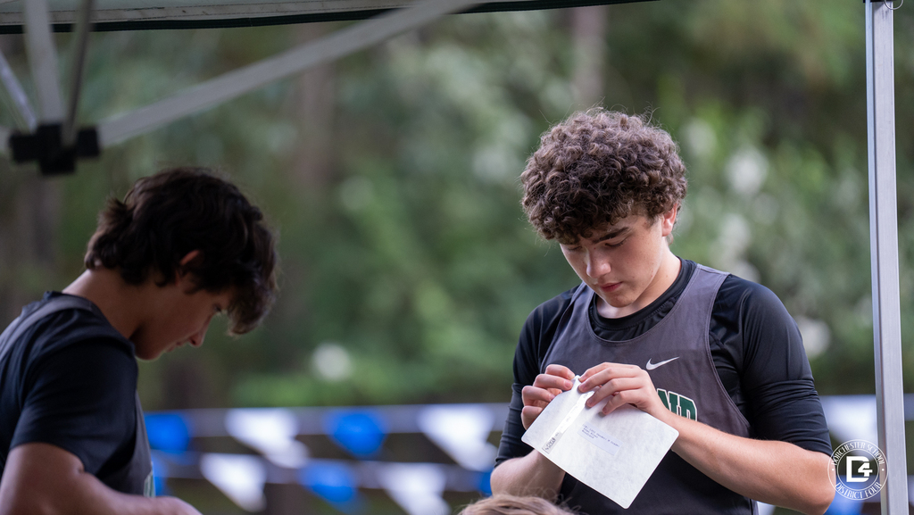 Two Woodland High School cross country runners prepare under a team tent at Wannamaker Park. A runner with curly hair, wearing a Woodland jersey over a black long sleeve shirt, peels the backing off his race bib.