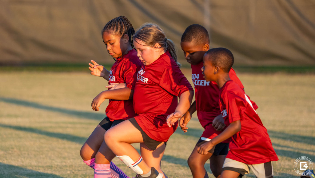 Four young players in red DD4 Soccer shirts run side by side, chasing the ball during a game at sunset.